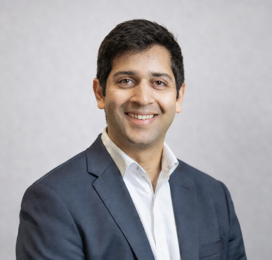 A man in a suit and white shirt is smiling in front of a wooden wall.