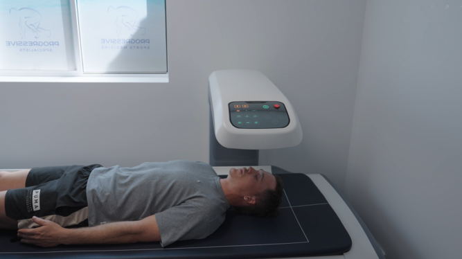 Person lying on a treatment table beside a medical device in a bright exam room