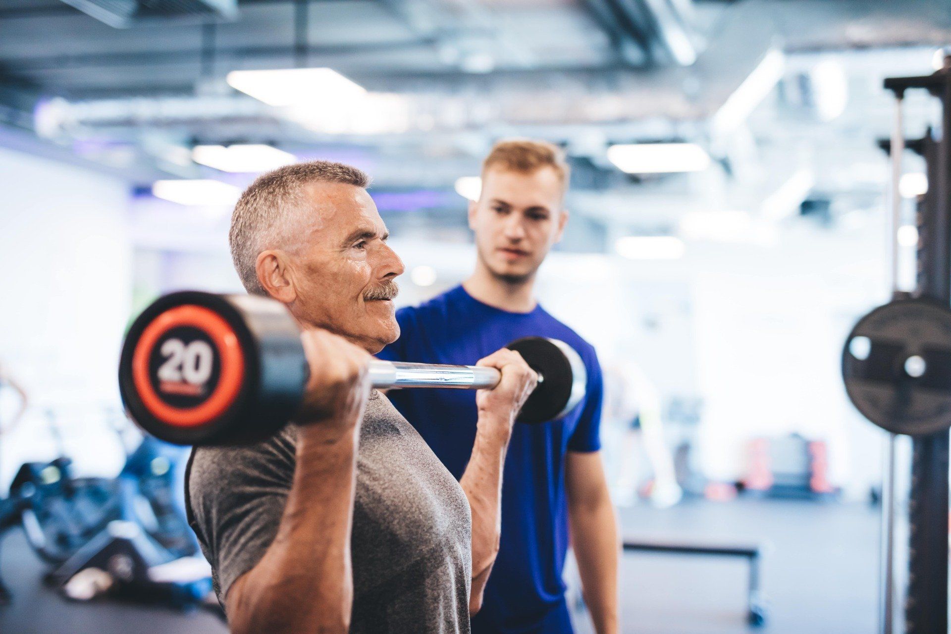 Man lifting a barbell in a gym while a trainer watches in the background