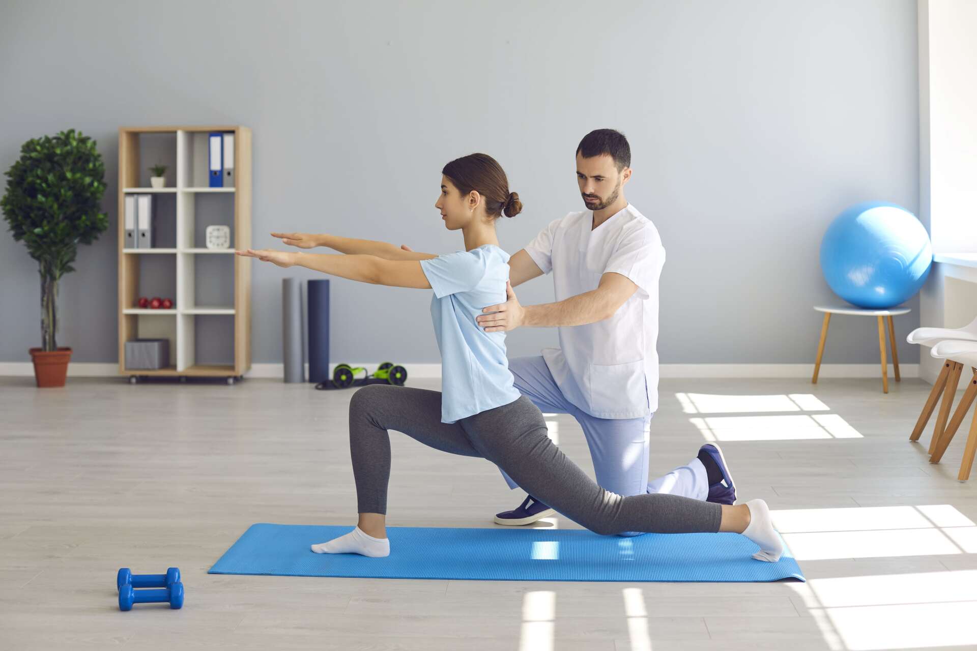 Two people practicing yoga in a bright room on blue mats, one assisting a lunge pose
