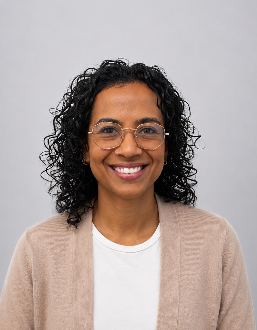 Smiling person with curly hair and glasses, wearing a beige cardigan over a white shirt against a gray background