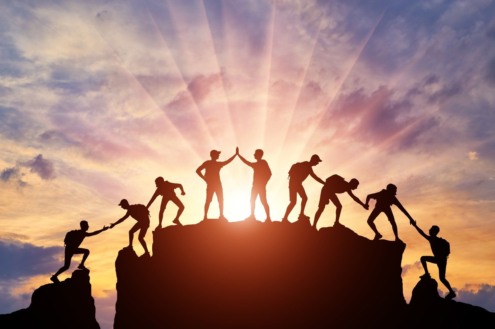 Silhouetted climbers helping each other atop a rocky peak at sunrise with dramatic orange sky
