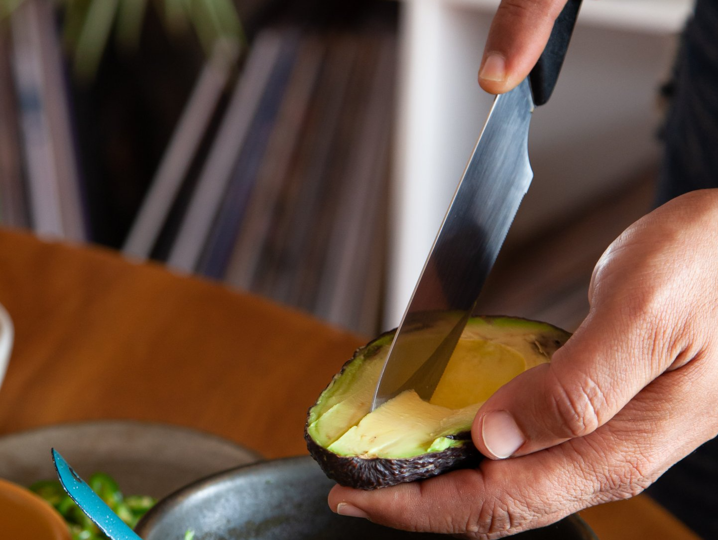 Hands slice avocado on a wooden table with a knife.