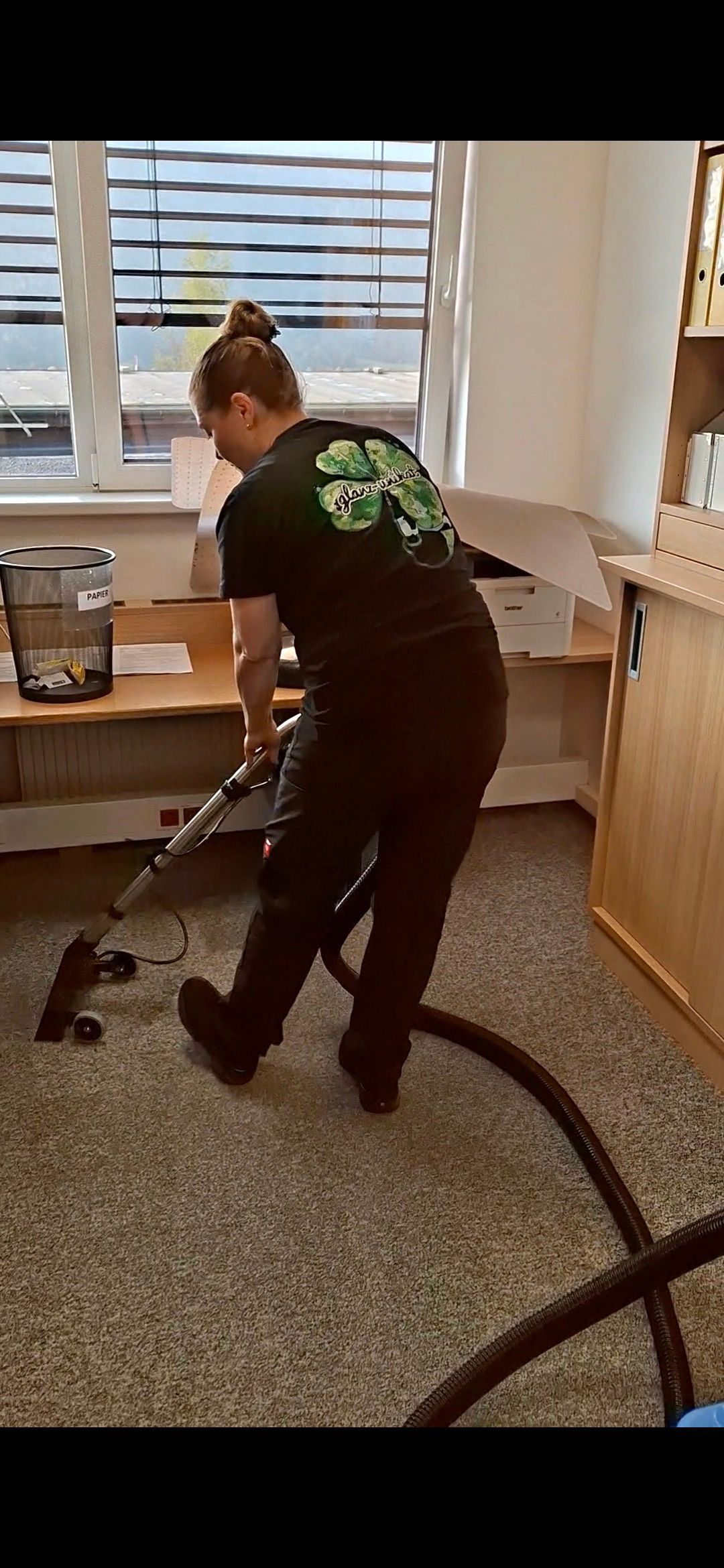 Eine Frau saugt Staub in einem mit Teppichboden ausgelegten Zimmer; im Bild sind ein Fenster,
