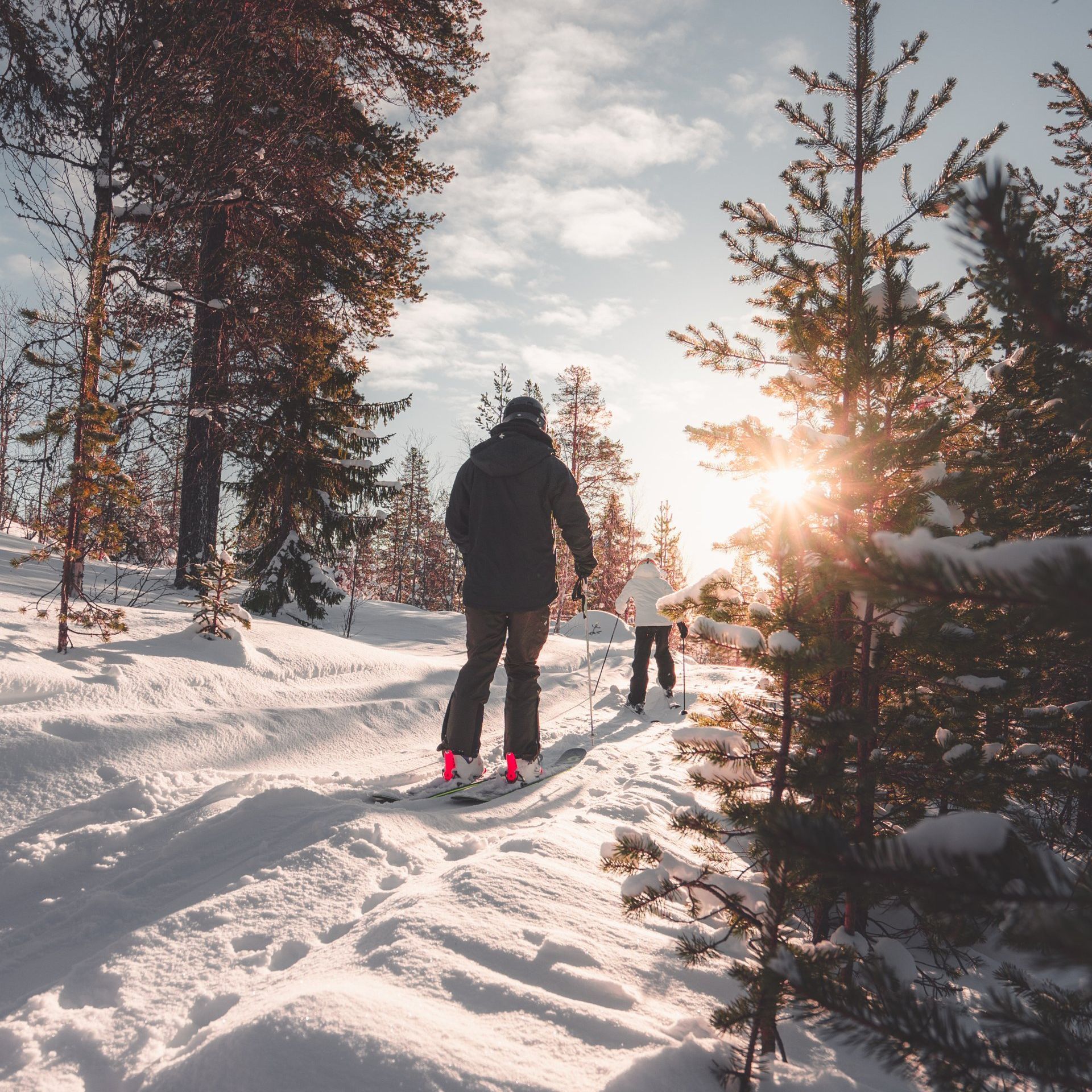 Two people skiing in the snow with the sun shining through the trees