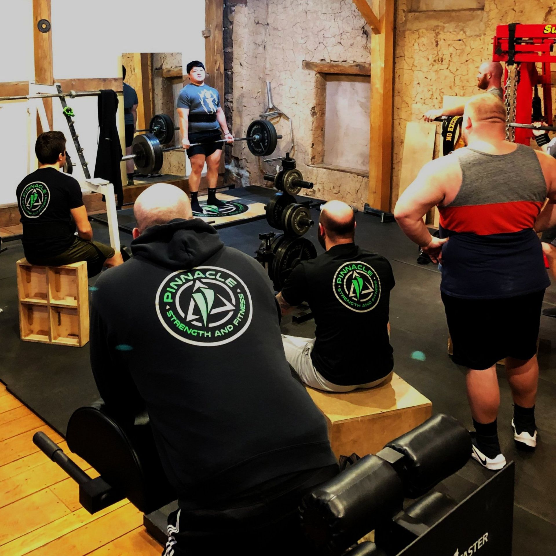 People seated in a gym watching a lifter perform a deadlift near weight racks.
