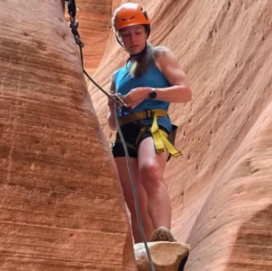 Climber in orange helmet rappels through a narrow sandstone canyon, holding a rope and wearing a harness.