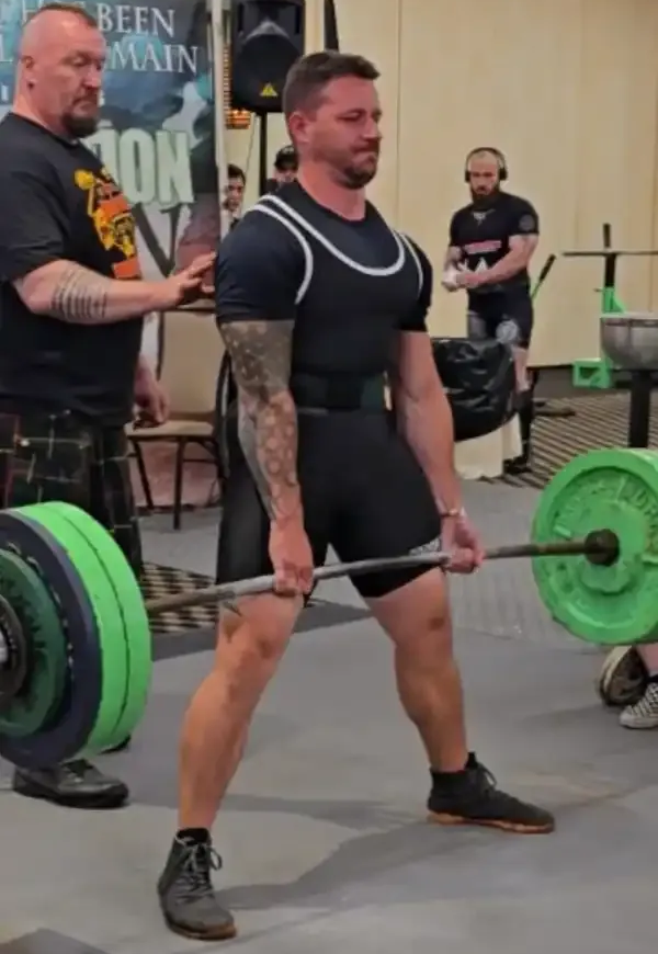 Weightlifter performing a deadlift in a gym, with green bumper plates and spotters in the background