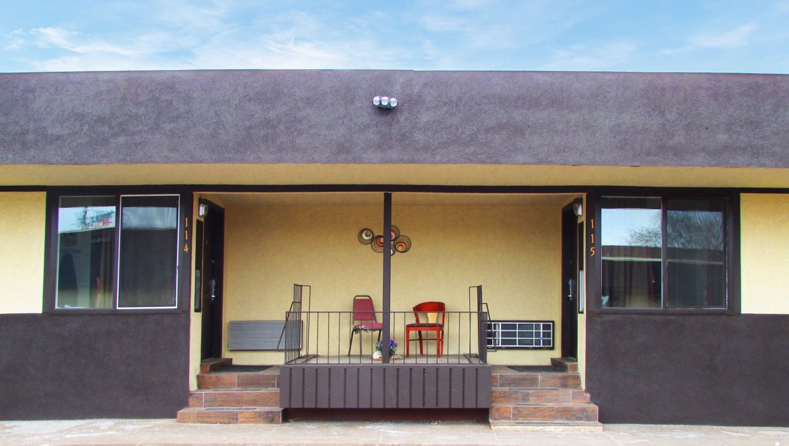 Exterior of a motel, beige walls, dark brown trim, small porch with two chairs and railing, under a cloudy blue sky.