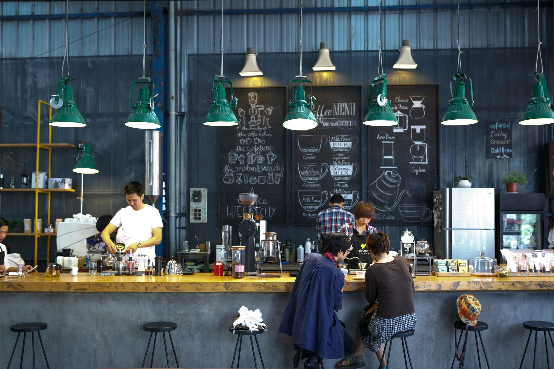 Coffee shop interior with bar, patrons, chalkboard menu, and green pendant lights.