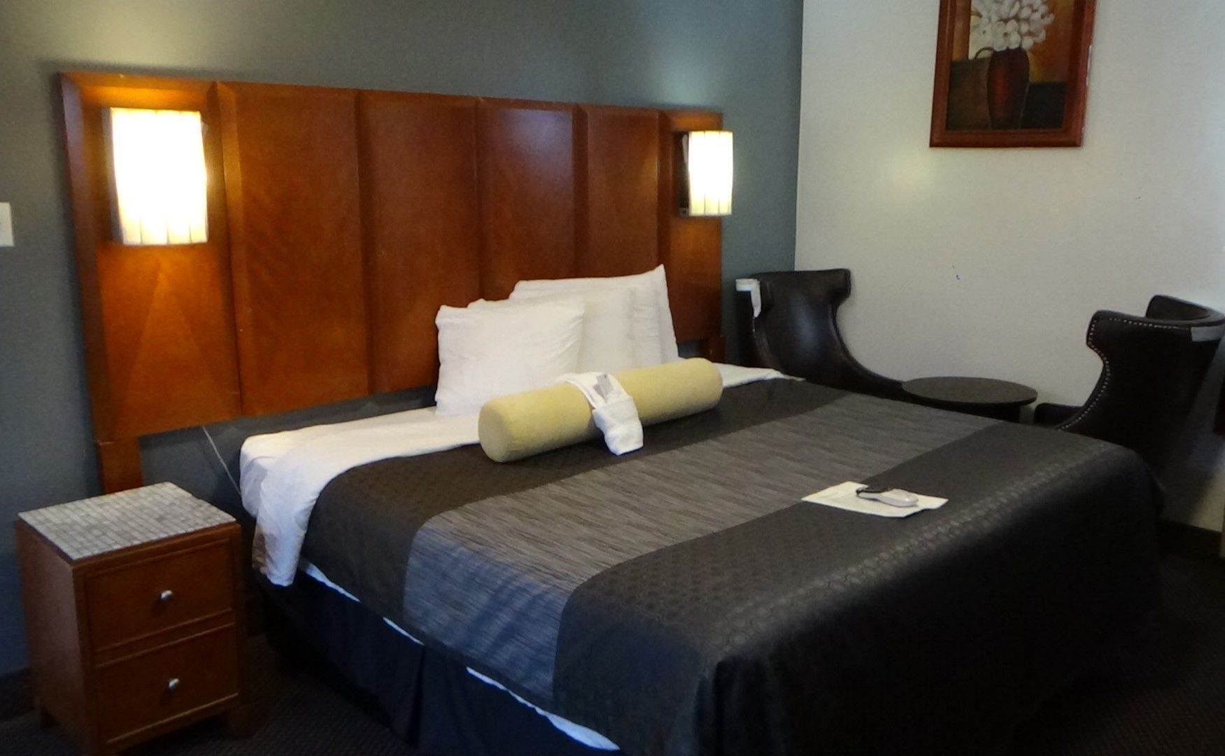 Hotel room with a large bed, dark wood headboard, two chairs, nightstand, and small artwork.