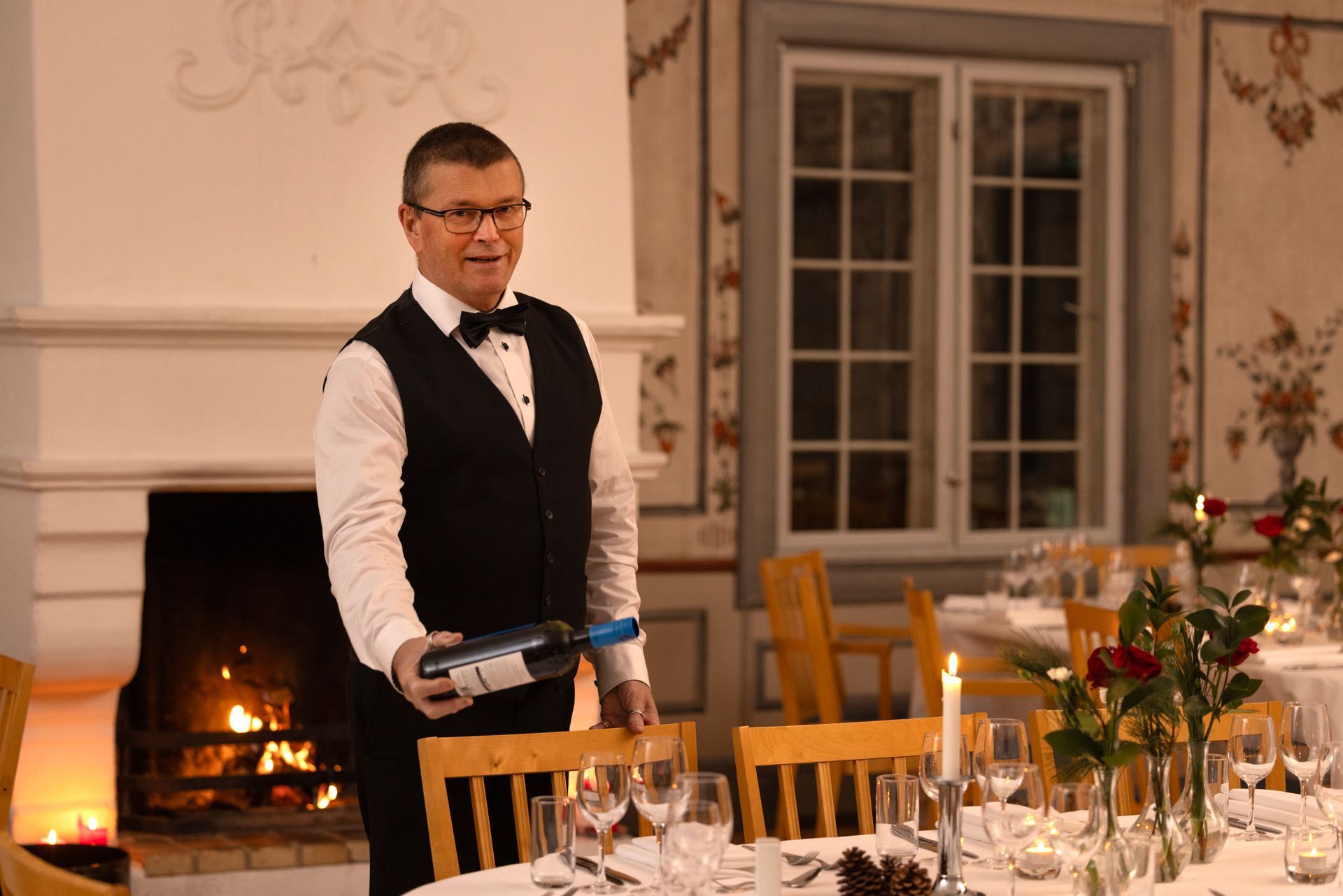 Kelner i svart vest som holder en vinflaske, står ved et dekket bord til en formell middag.