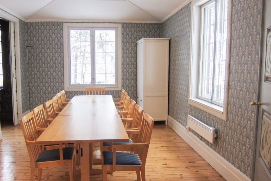 Spisestue med langt trebord, stoler, tapet, vinduer og hvitt skap.