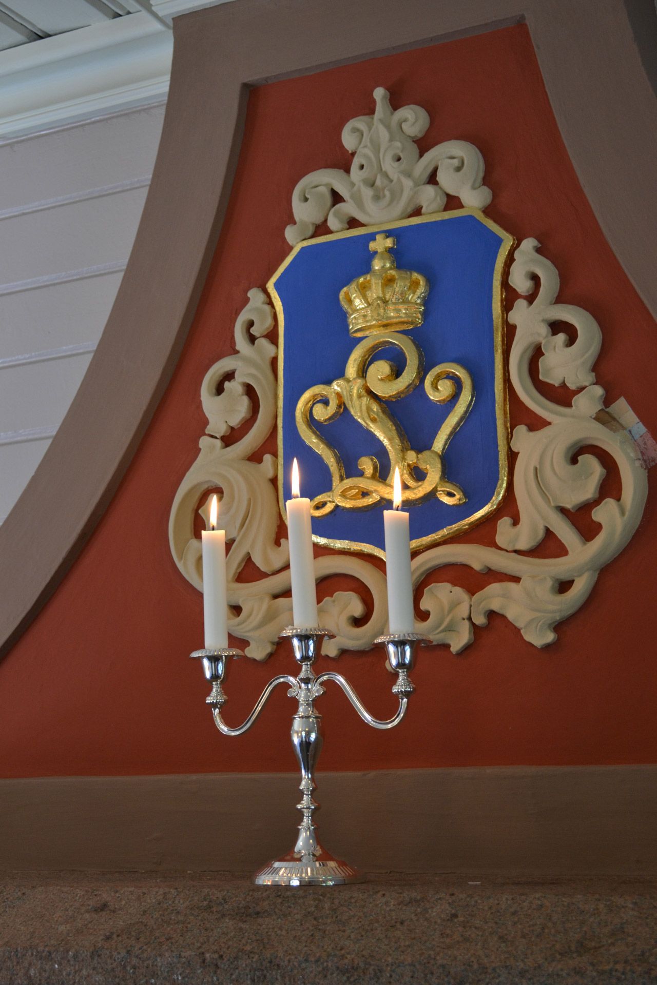 Sølvlysesnegl med tente lys foran et blått skjold med gyllen krone og monogram på en rød vegg.