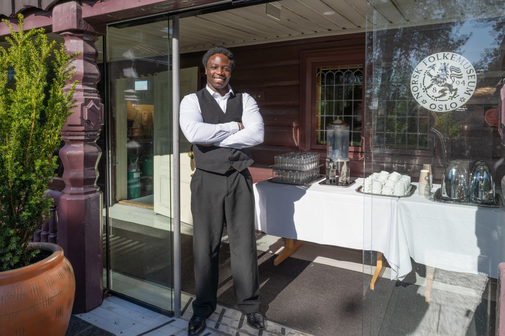 En smilende person står foran døråpningen til en restaurant med armene i kors, ved siden av et bord med mat.