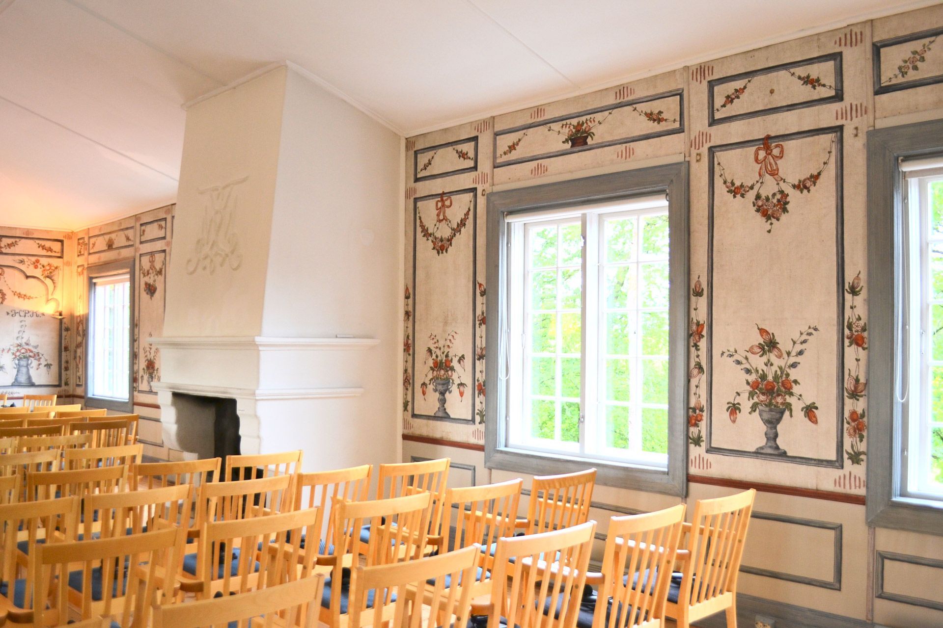 Innvendig rom med rader med trestoler, blomsterdekorasjon på veggen og vinduer.