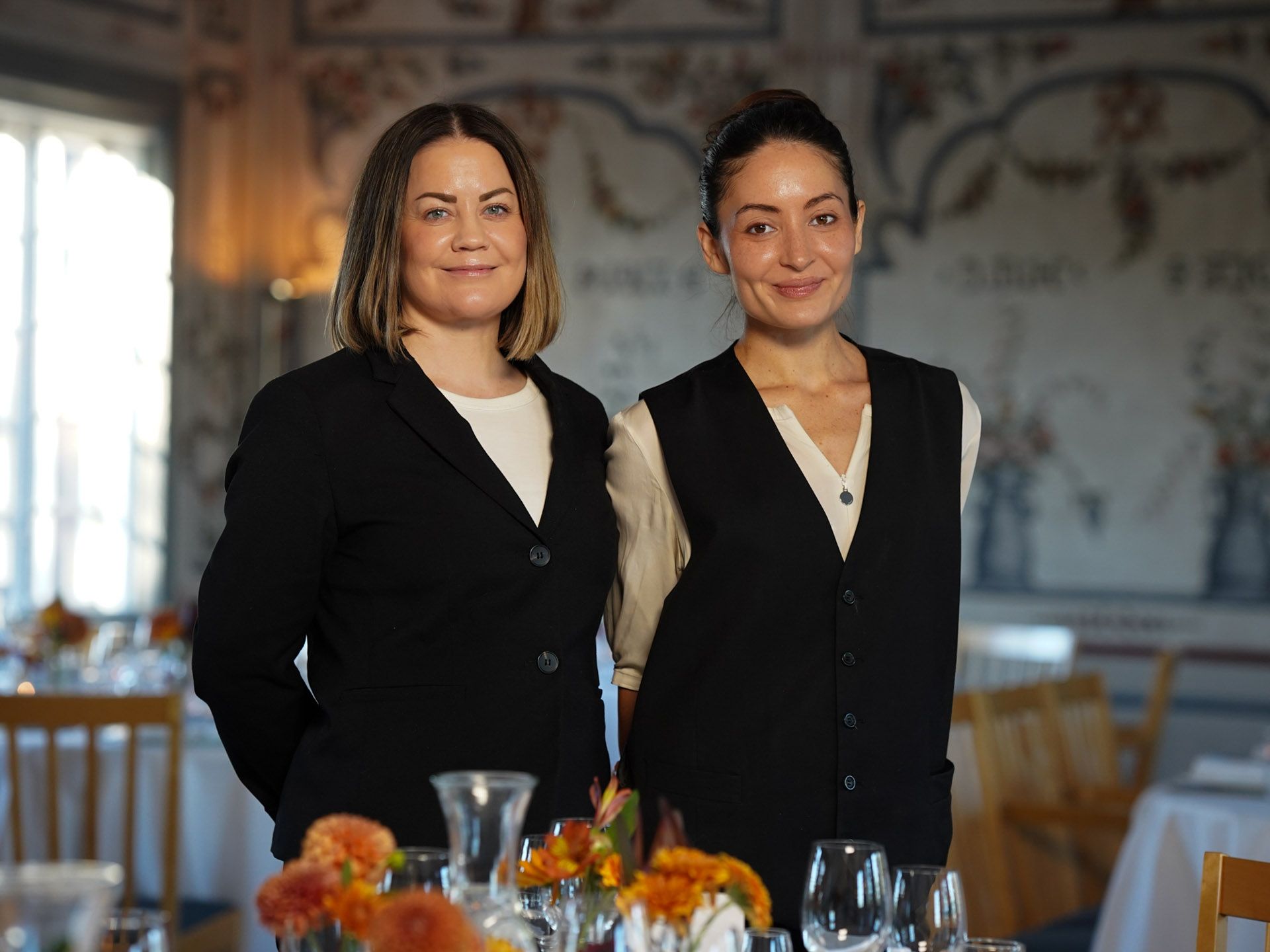 To kvinner står ved et spisebord, begge i svarte vester og hvite topper, og smiler. Formell restaurantsetting.