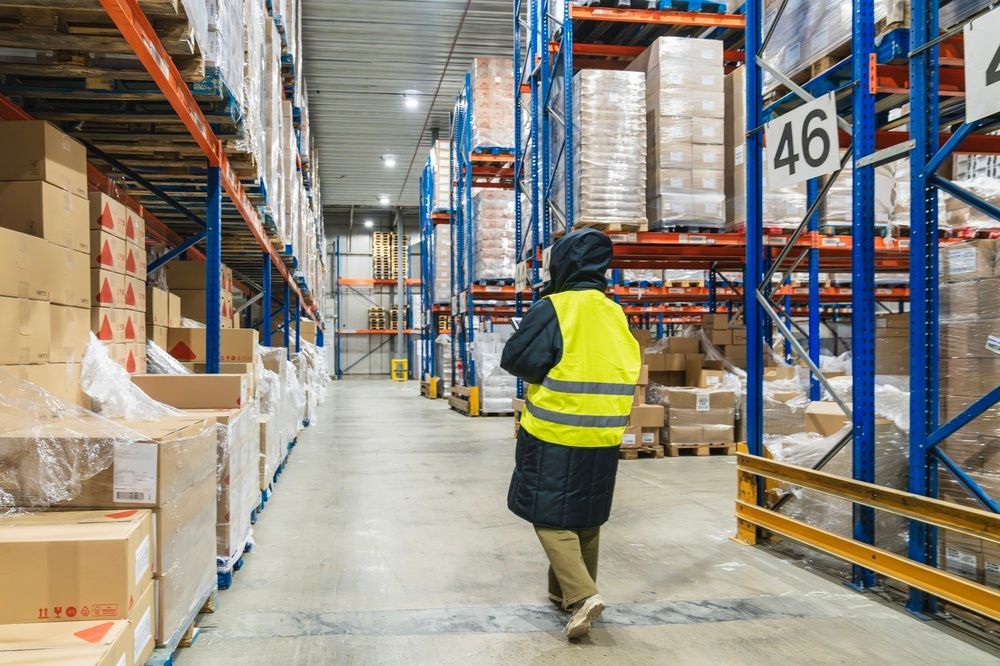 Warehouse worker in a yellow safety vest walking through a stocked warehouse aisle
