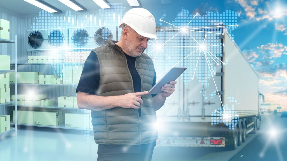 Worker in a hard hat using a tablet in a bright warehouse with a truck in the background