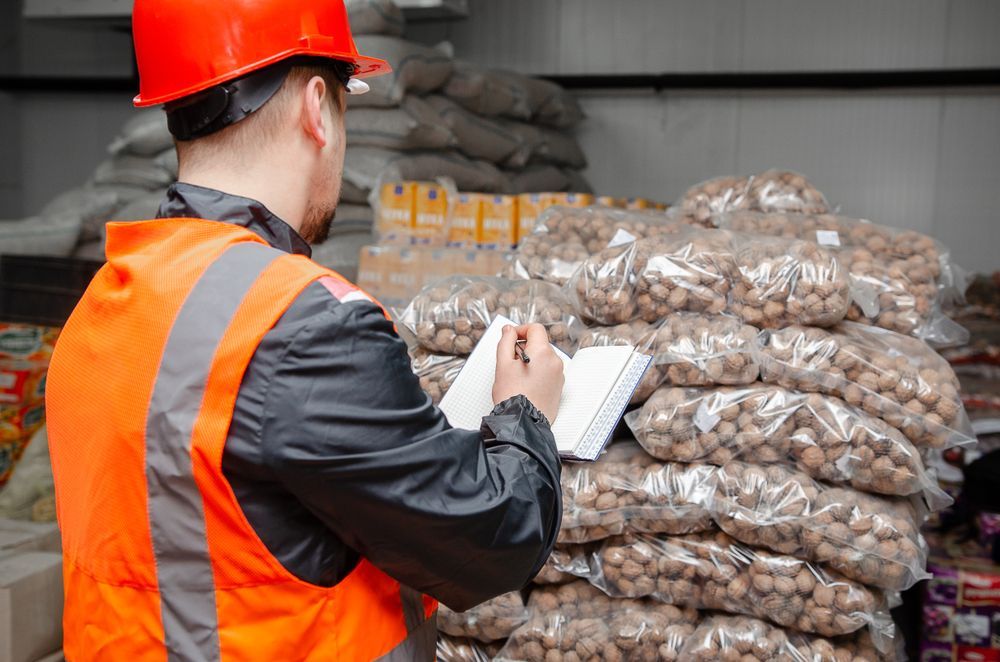 Worker in orange safety vest and hard hat taking notes beside stacked bags in a warehouse.