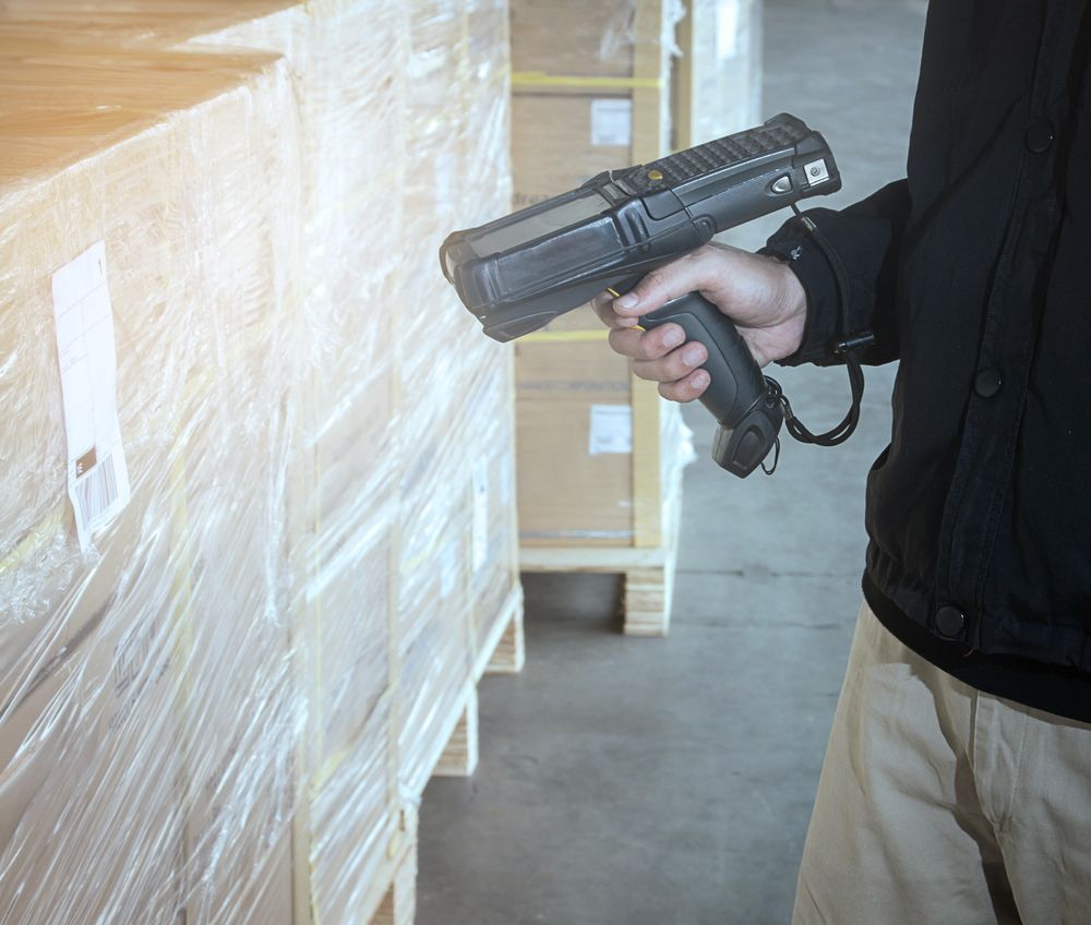 Person holding a handheld scanner near stacked wooden pallets in a warehouse.