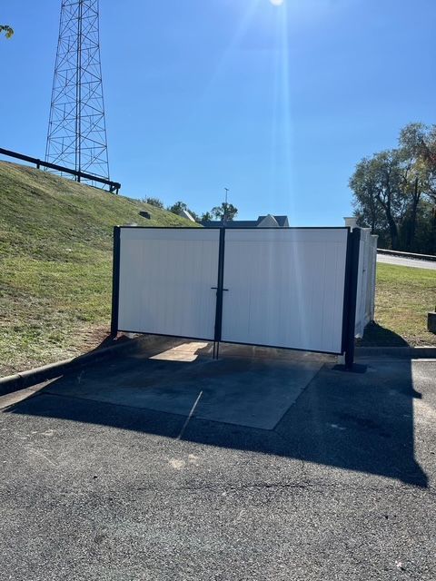 White-walled enclosure with black posts and double doors on asphalt, set against a grassy hill and blue sky.