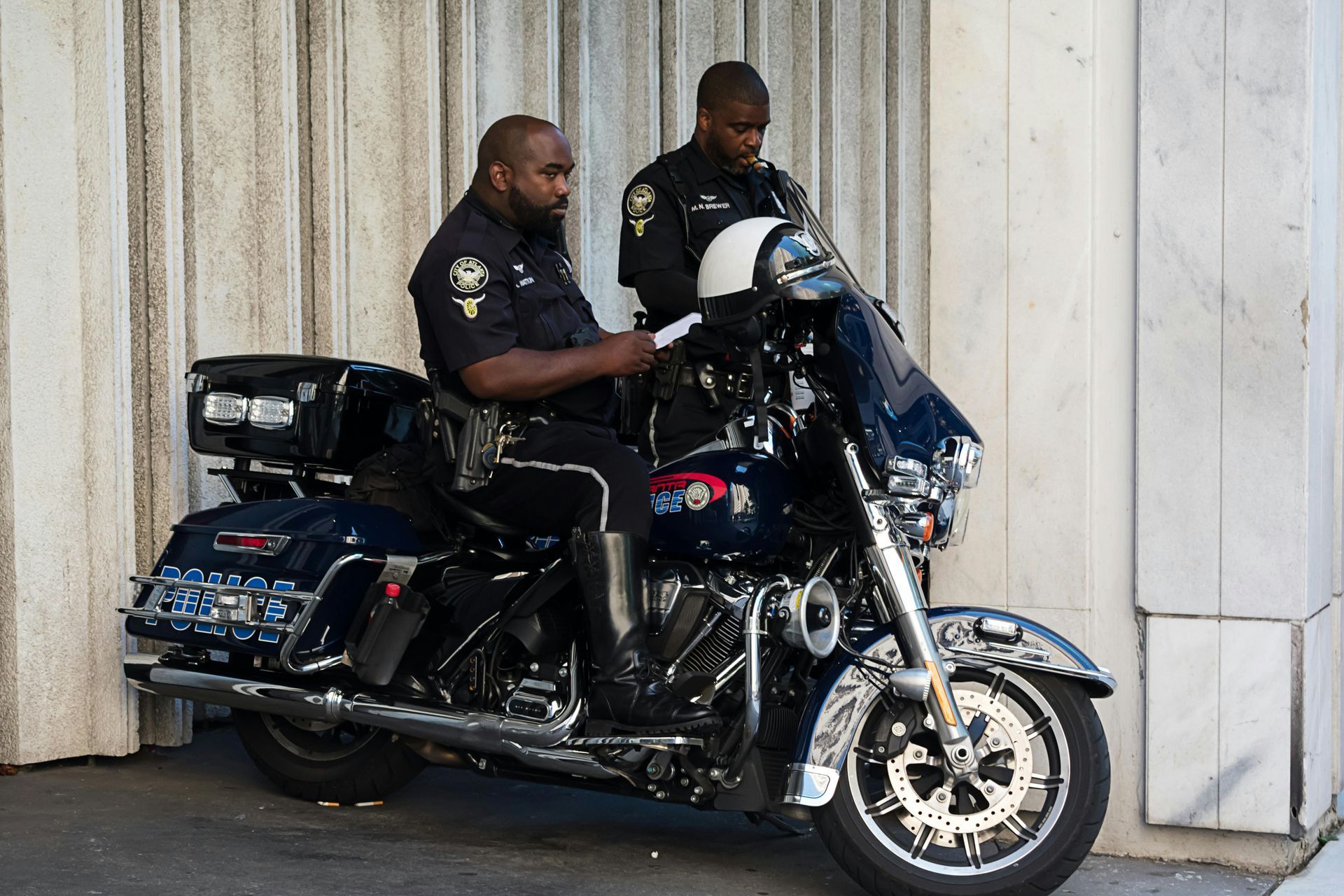 Two police officers beside a blue motorcycle, reviewing paperwork near a building.