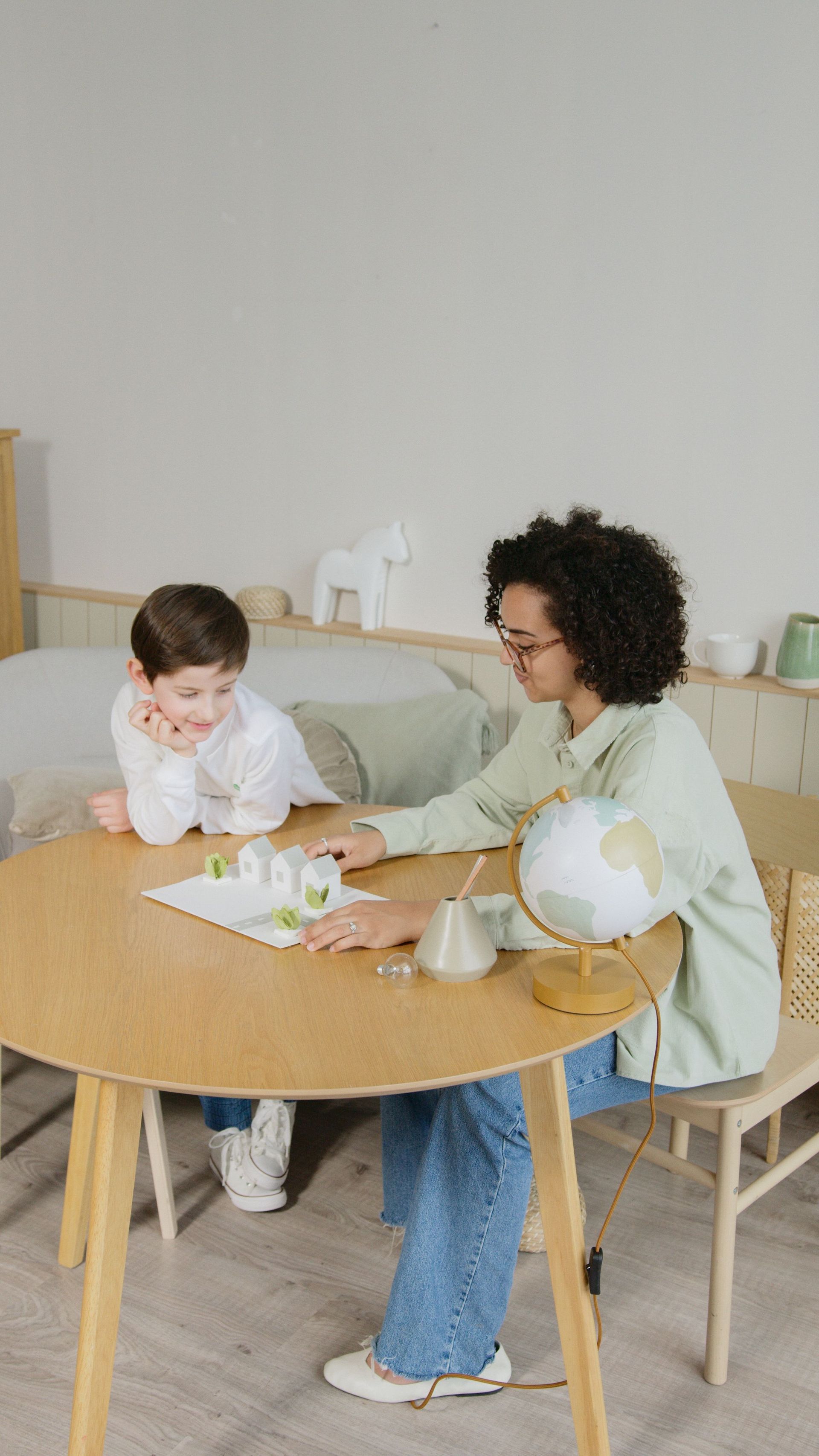 Two people sitting at a wooden table, writing on paper in a bright room with crafts supplies.