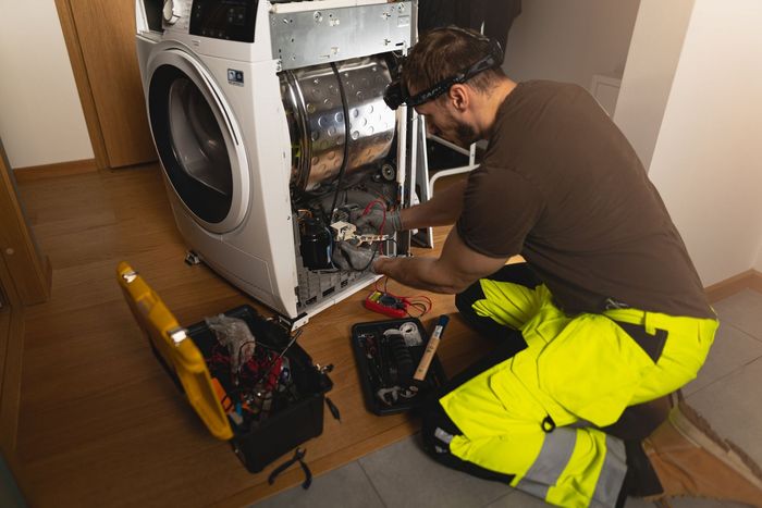 A technician in yellow work pants repairs a washing machine, tools and equipment are around the machine.