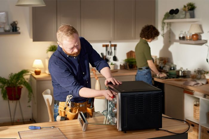 Man repairing an oven in a kitchen, a woman in the background near a sink.
