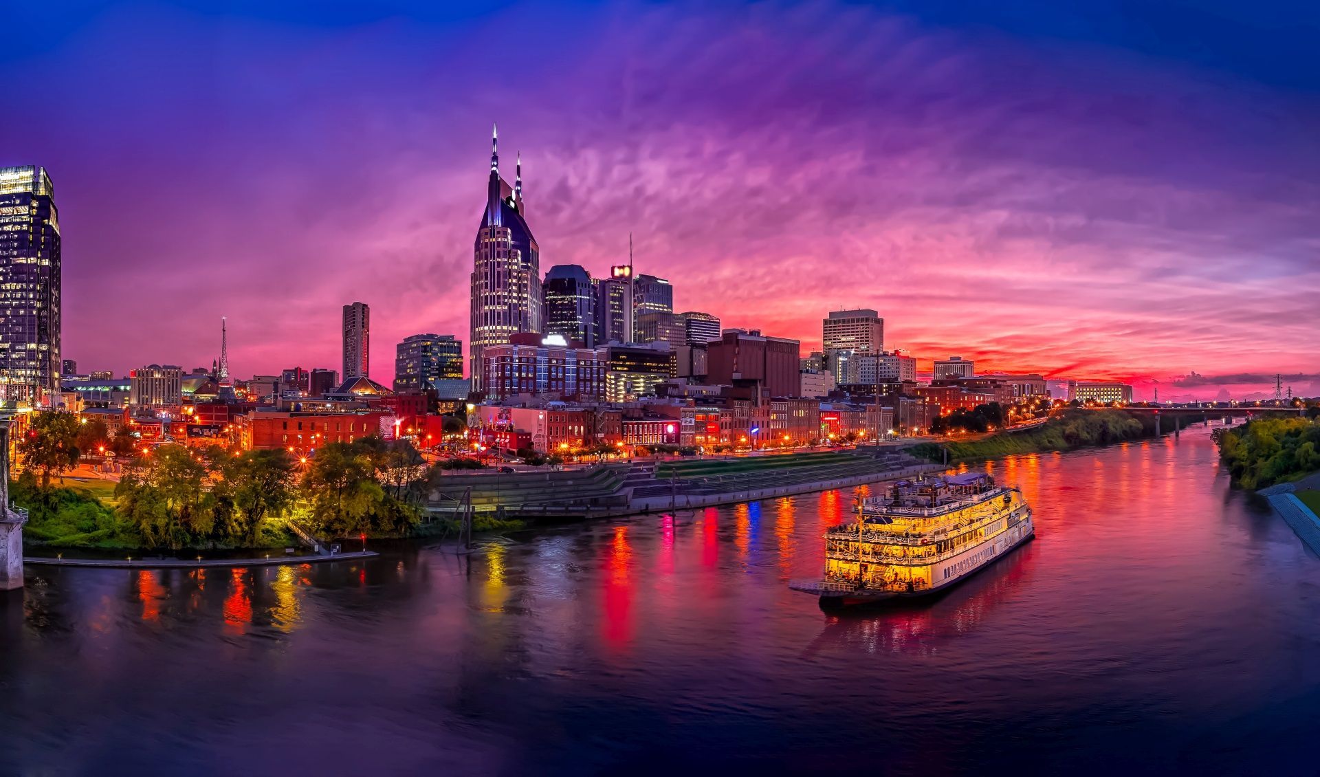 A boat is floating on a river in front of a city skyline at sunset.
