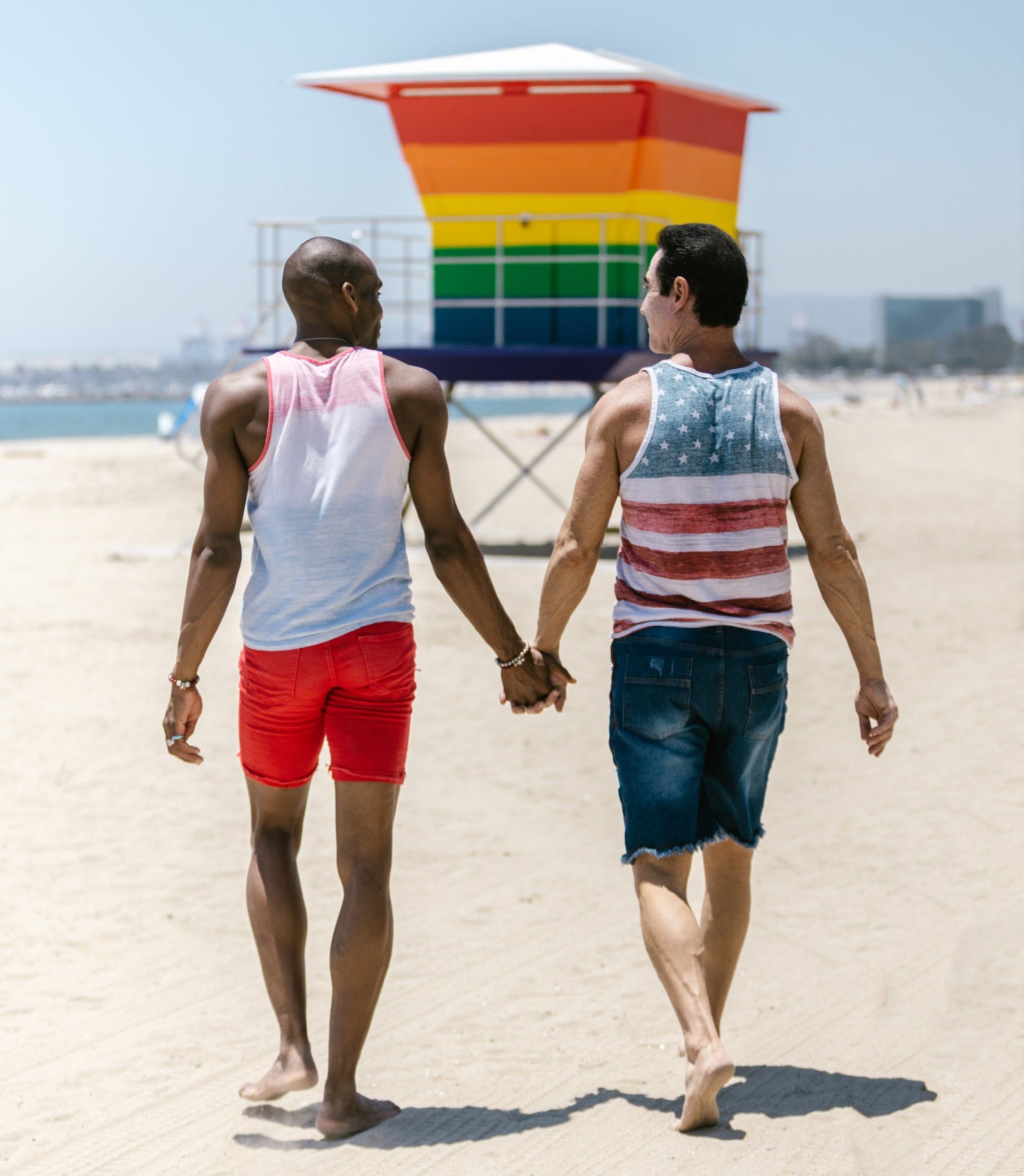 Two men are walking on the beach holding hands in front of a lifeguard tower