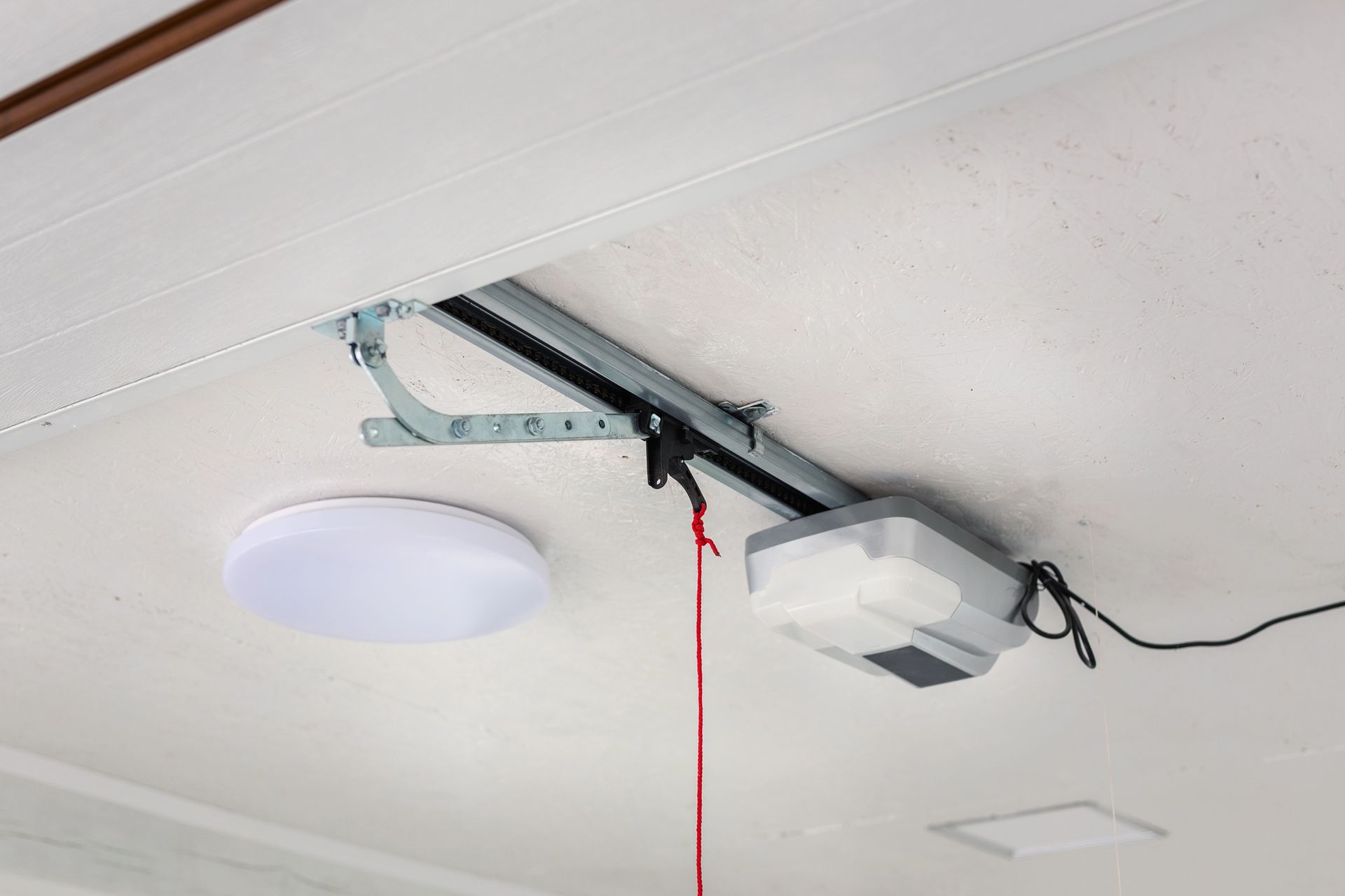 A ceiling-mounted garage door opener with a metal rail, an arm, and a red emergency release cord.