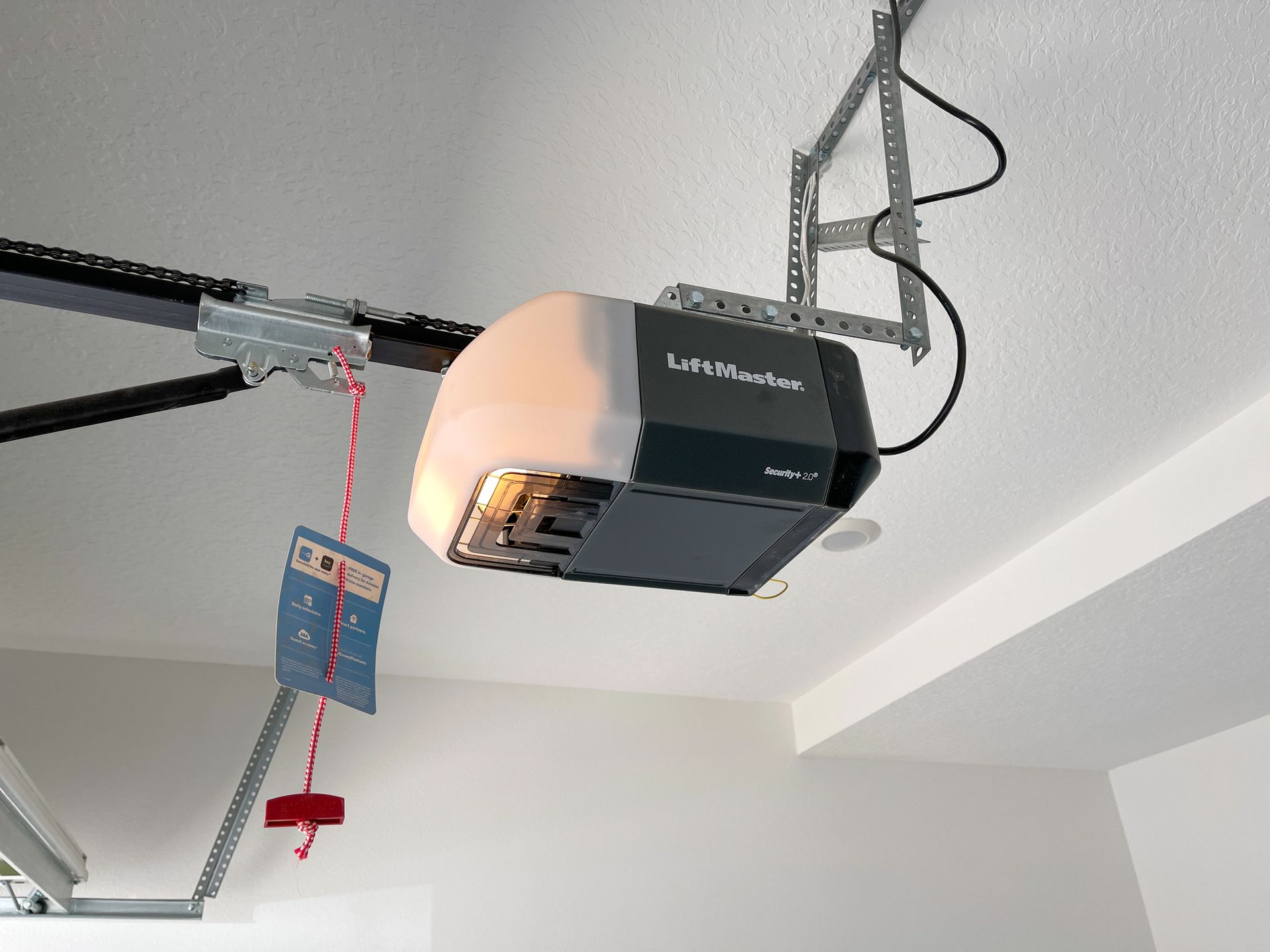 A black and white LiftMaster garage door opener mounted to a ceiling with a red manual release rope hanging down.