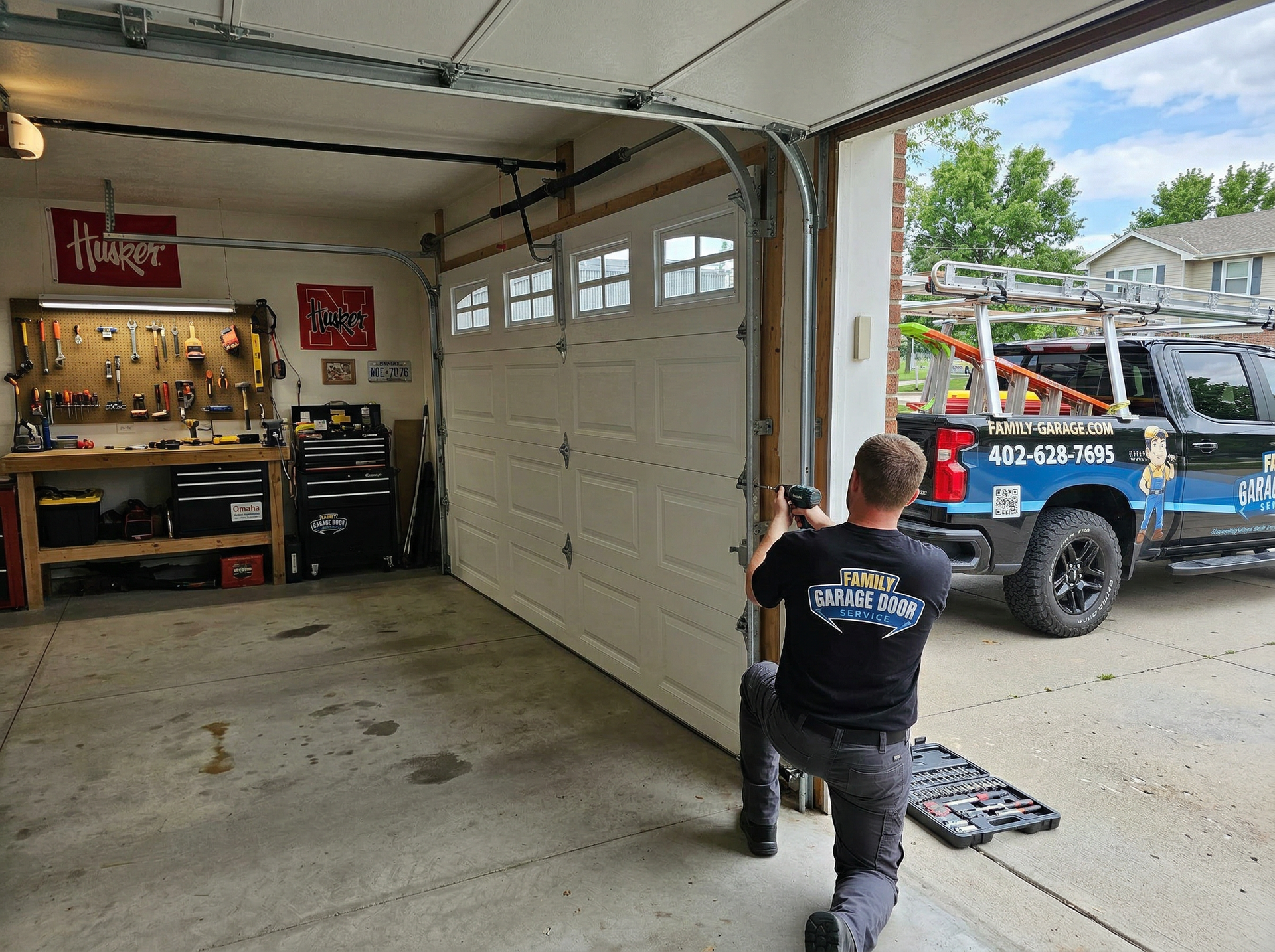 A technician wearing a work shirt kneels in a garage, using a tool to repair the tracks of a white sectional garage door.