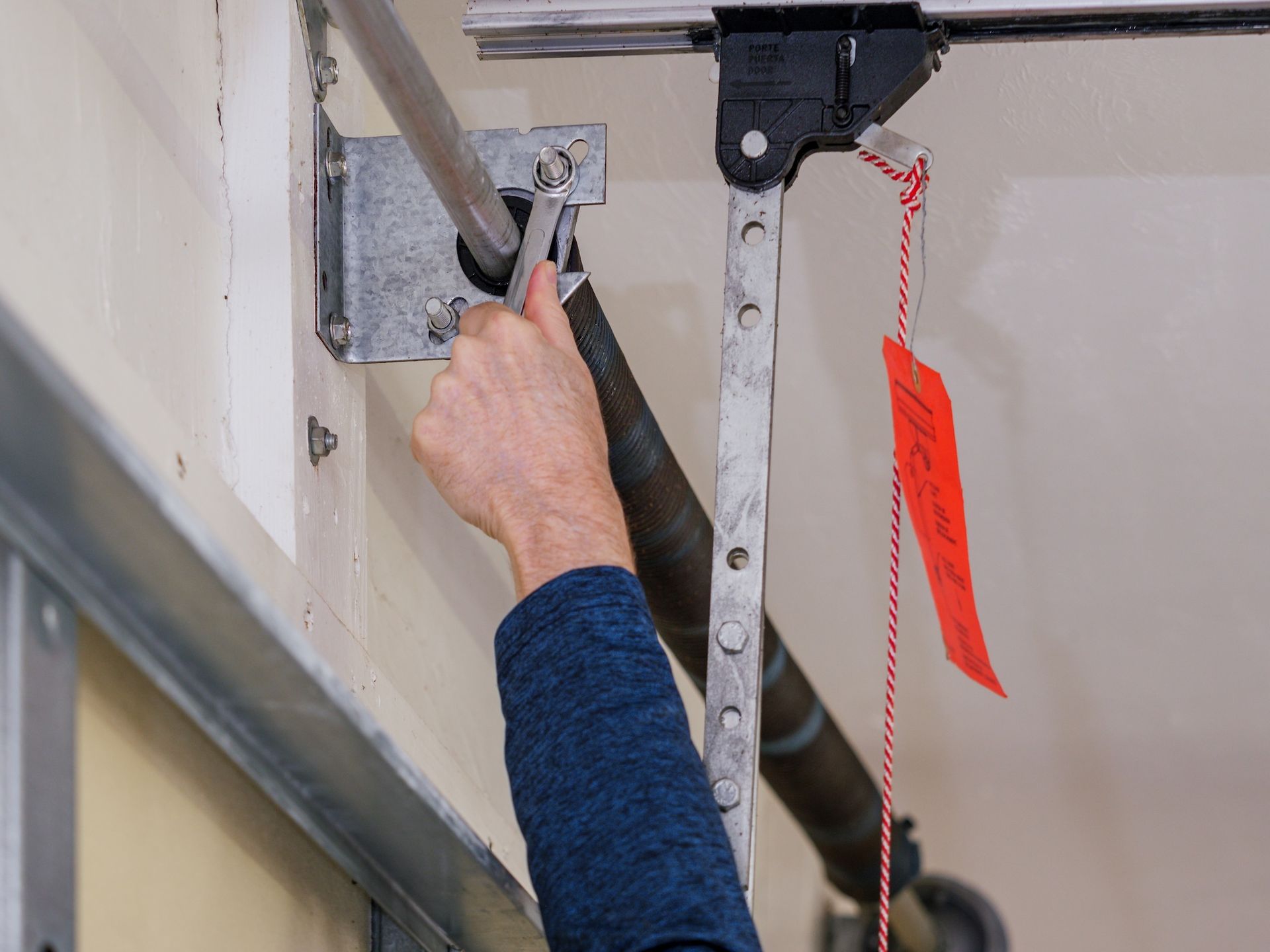 A person uses a wrench to adjust a garage door torsion spring system near a red emergency release handle.