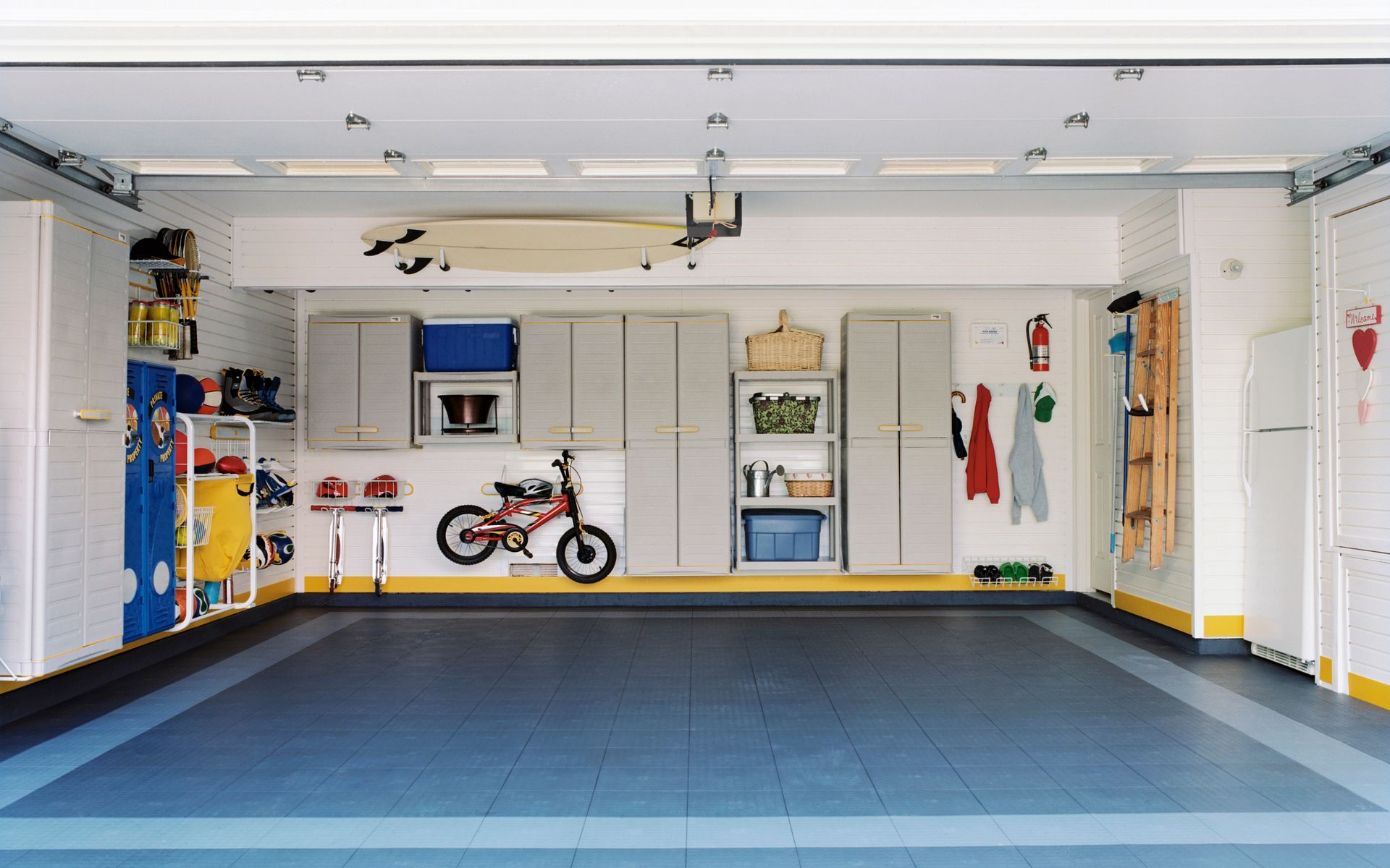 A well-organized garage with blue and grey floor tiles, storage cabinets, a bicycle, and a surfboard mounted overhead.