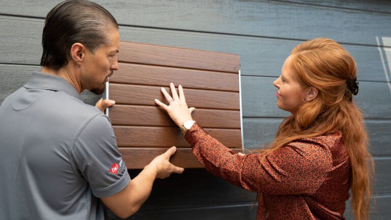 Two people comparing wood siding samples against a gray exterior wall