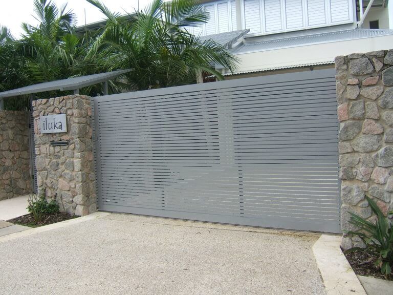 Gray metal driveway gate beside stone pillars and tropical plants, with a house number plaque on the left.