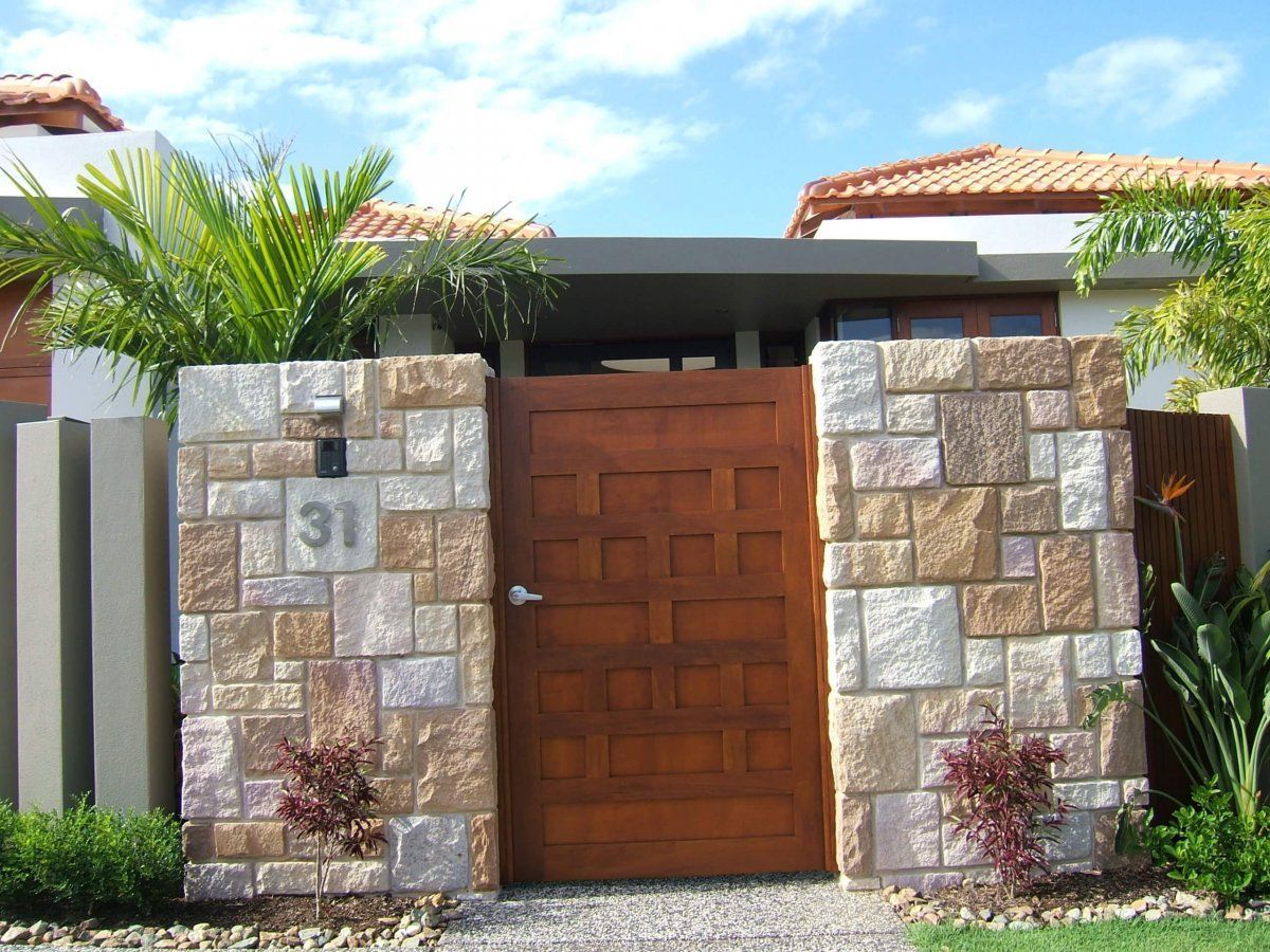 Modern house entrance with wooden gate, stone walls, palm trees, and a blue sky.