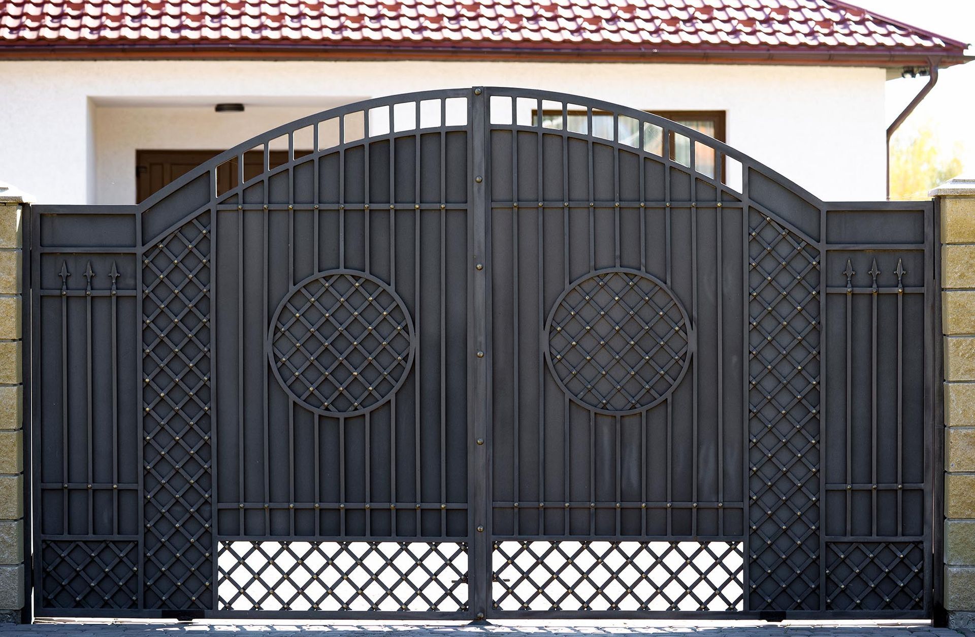 Black metal double driveway gate in front of a house with a tiled roof.