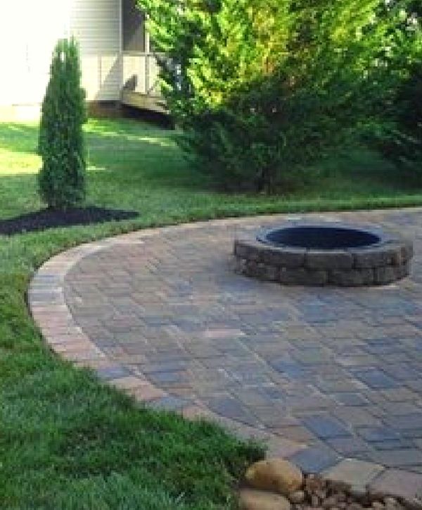 A backyard stone patio with a circular fire pit surrounded by a grassy lawn, shrubs, and trees.