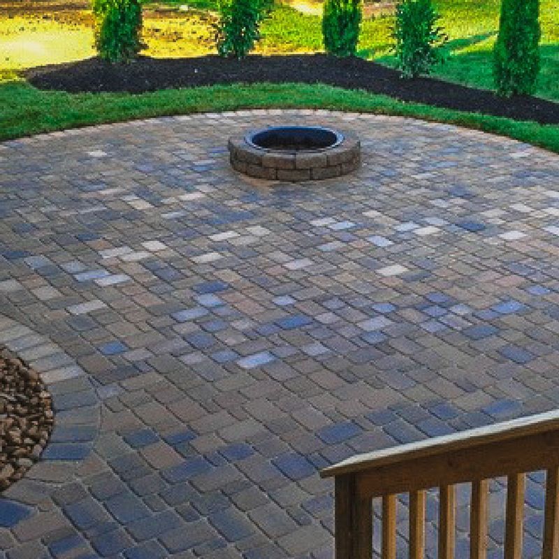 A circular paver patio with a stone fire pit in the center, bordered by small evergreen trees and a wooden deck railing.