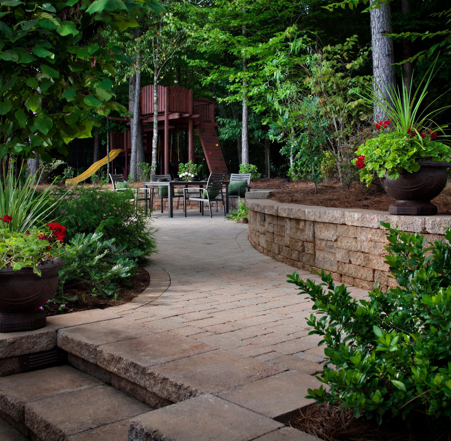 A stone walkway leads to a patio with a wooden play structure, surrounded by trees, potted plants, and a retaining wall.