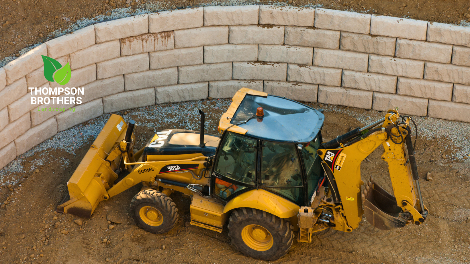 A yellow backhoe loader sits inside a curved, beige concrete block retaining wall under construction.
