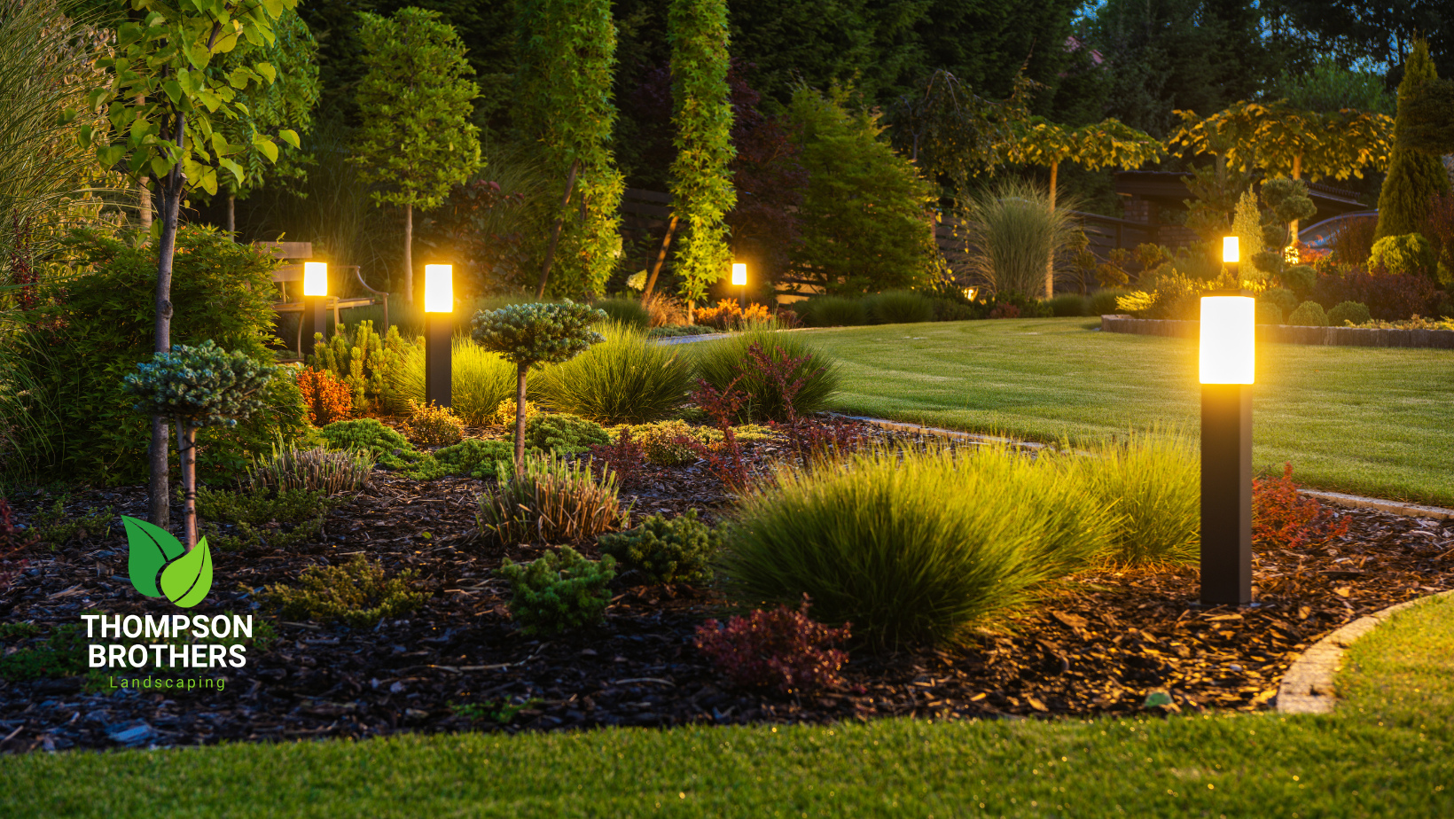 A lush garden at dusk featuring glowing modern pathway lights, diverse greenery, and dark mulch beds.