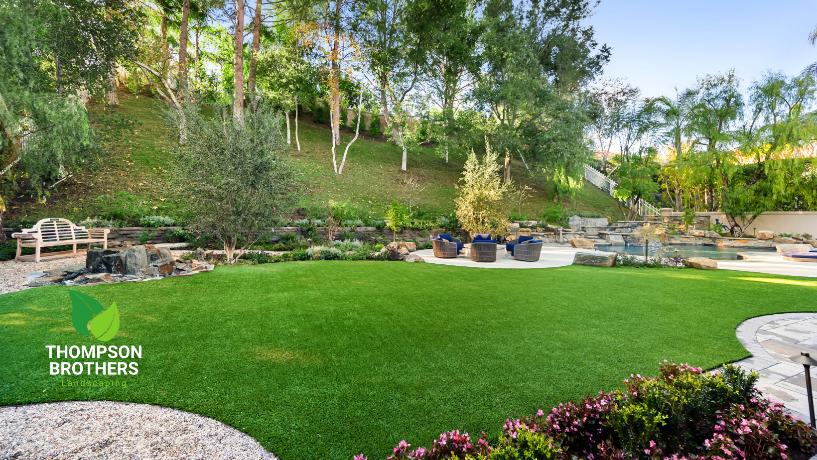 Lush green backyard with artificial turf, stone patio, landscaping, and a hillside. Thompson Brothers logo visible.