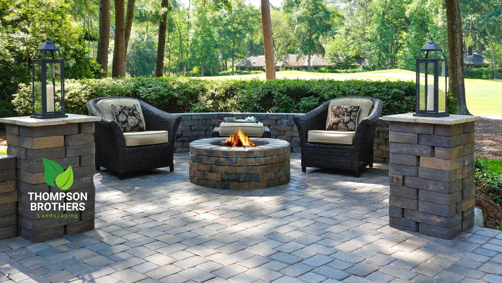 Patio with two wicker chairs, a circular stone fire pit, and stone pillars with lanterns in a backyard setting.