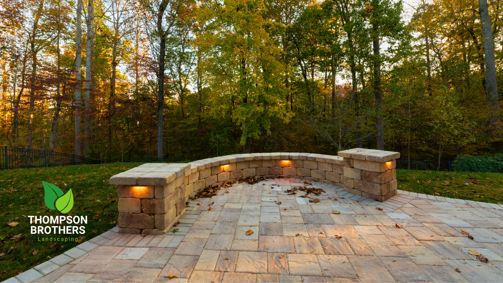 Curved stone garden wall with integrated lighting on a paved patio, set against a backdrop of autumn trees.