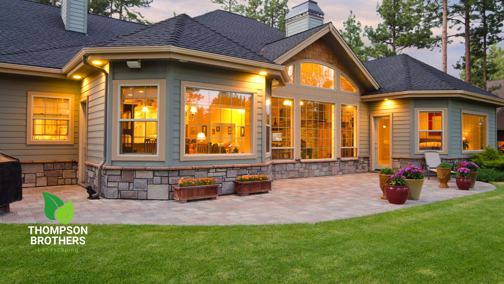 Back of a house with lit windows, stone accents, and patio with flower pots; green grass.